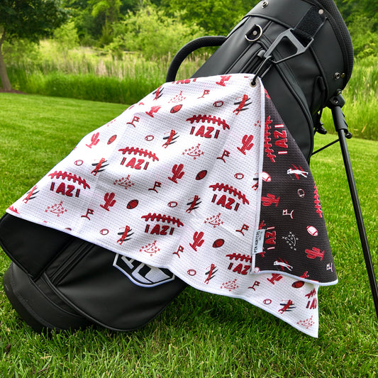 Arizona Cardinals white golf towel hanging on a black golf bag with red AZIfootball graphics, cactus icons, golfers, footballs, playbook, and black reverse side.