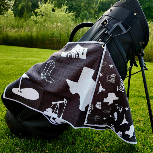 Close up of a black Texas themed golf towel with white Texas state, longhorn, oil pump, cowboy boot, Alamo, and golf graphics.