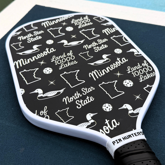 Black and white Minnesota pickleball paddle close-up with loon icons, state outlines, North Star State text, and Land of 10,000 Lakes graphics.