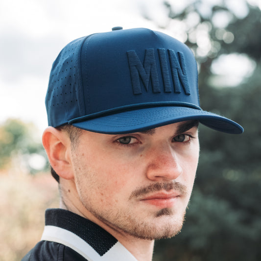 Blue tonal MIN performance golf hat with raised matching embroidery, flat brim, rope detail, and clean front view on model.