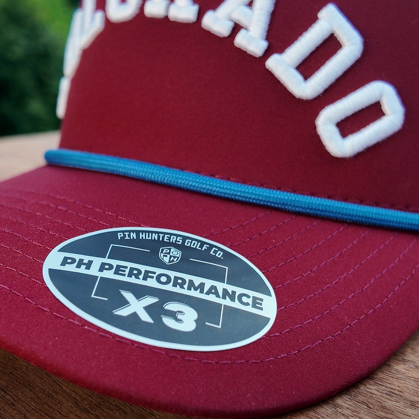 Close up of the Colorado Avalanche performance golf hat showing maroon brim, blue rope, raised white Colorado embroidery, and PH Performance X3 sticker.