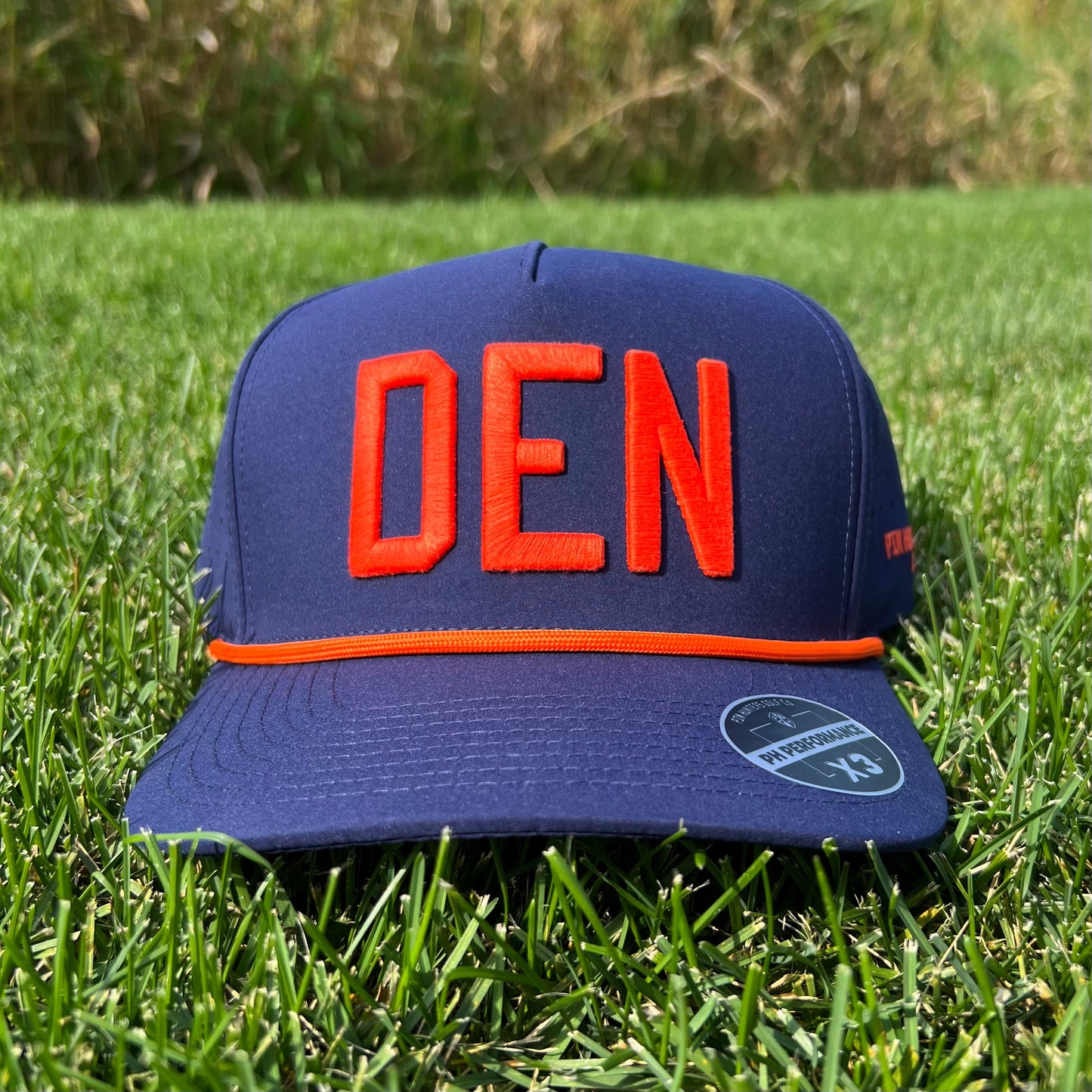 Navy Denver performance golf hat on grass with bright orange raised DEN embroidery, orange rope, flat brim, and PH Performance X3 sticker.