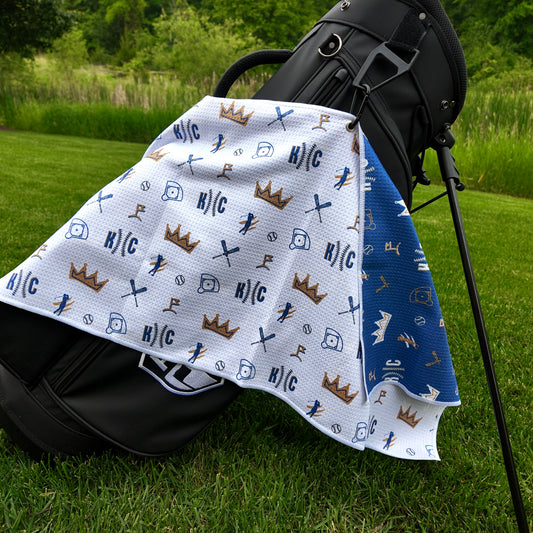 Kansas City Royals white golf towel hanging on a black golf bag with KC text, crown graphics, baseballs, golfers, golf flags, and blue reverse side.