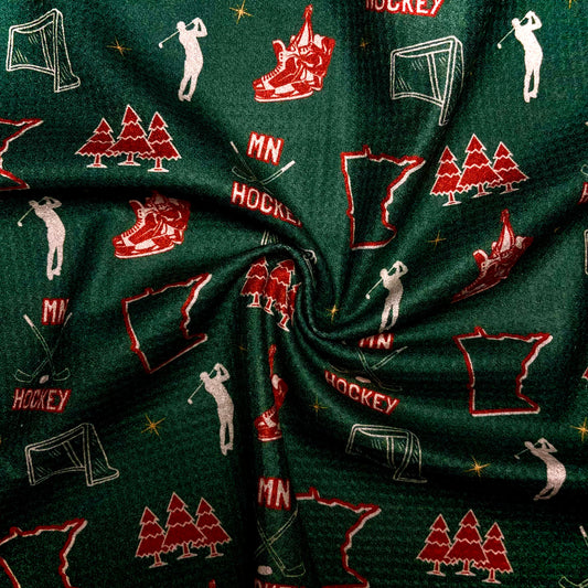 Close up of a green Minnesota Wild inspired hockey golf towel showing MN hockey text, red skates, hockey nets, Minnesota outlines, pine trees, golfers, and waffle microfiber texture.
