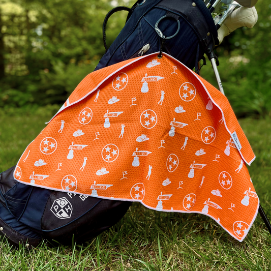 Orange Tennessee TriStar golf towel hanging from a golf bag, showing the orange back pattern with Tennessee TriStar logos, Tennessee text, golf flags, golfers, and golf icons.