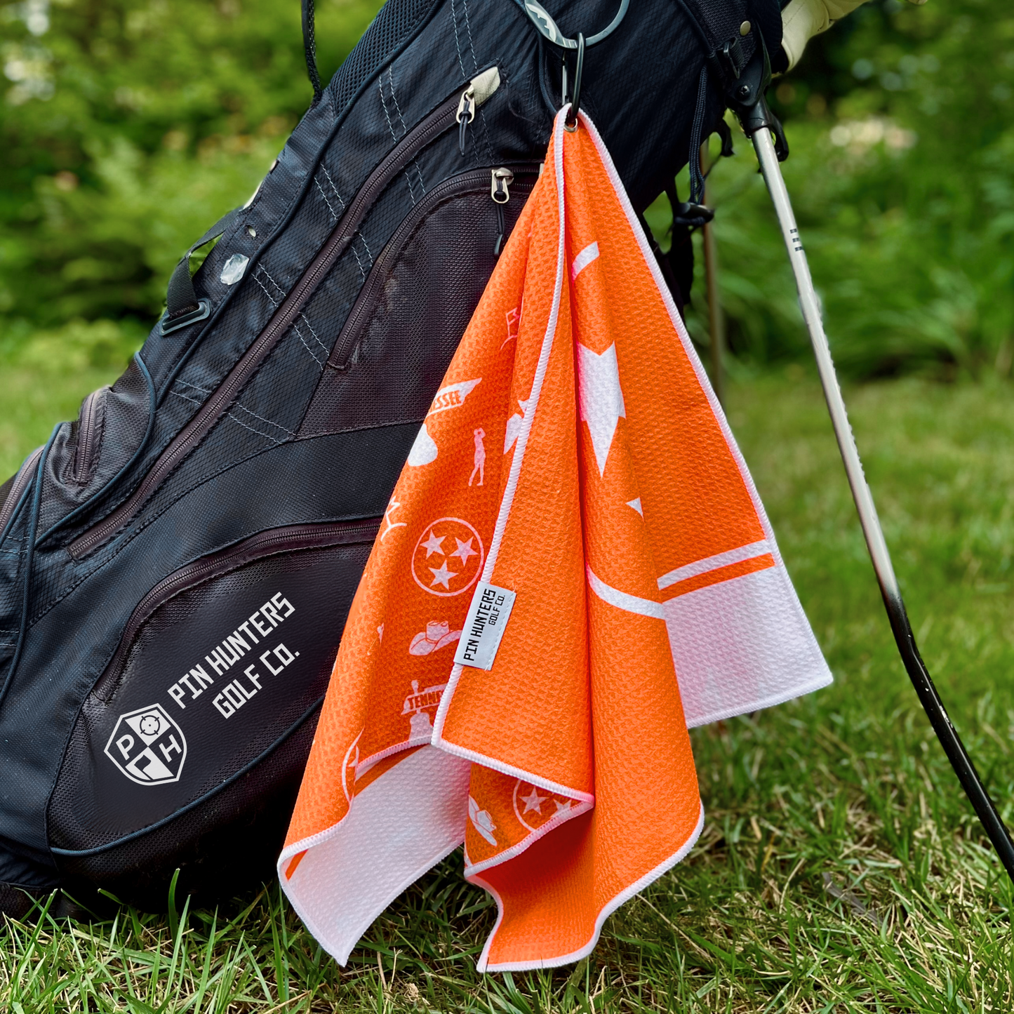 Orange Tennessee TriStar golf towel hanging from a navy golf bag, showing the folded towel, Pin Hunters label, and orange Tennessee themed pattern.