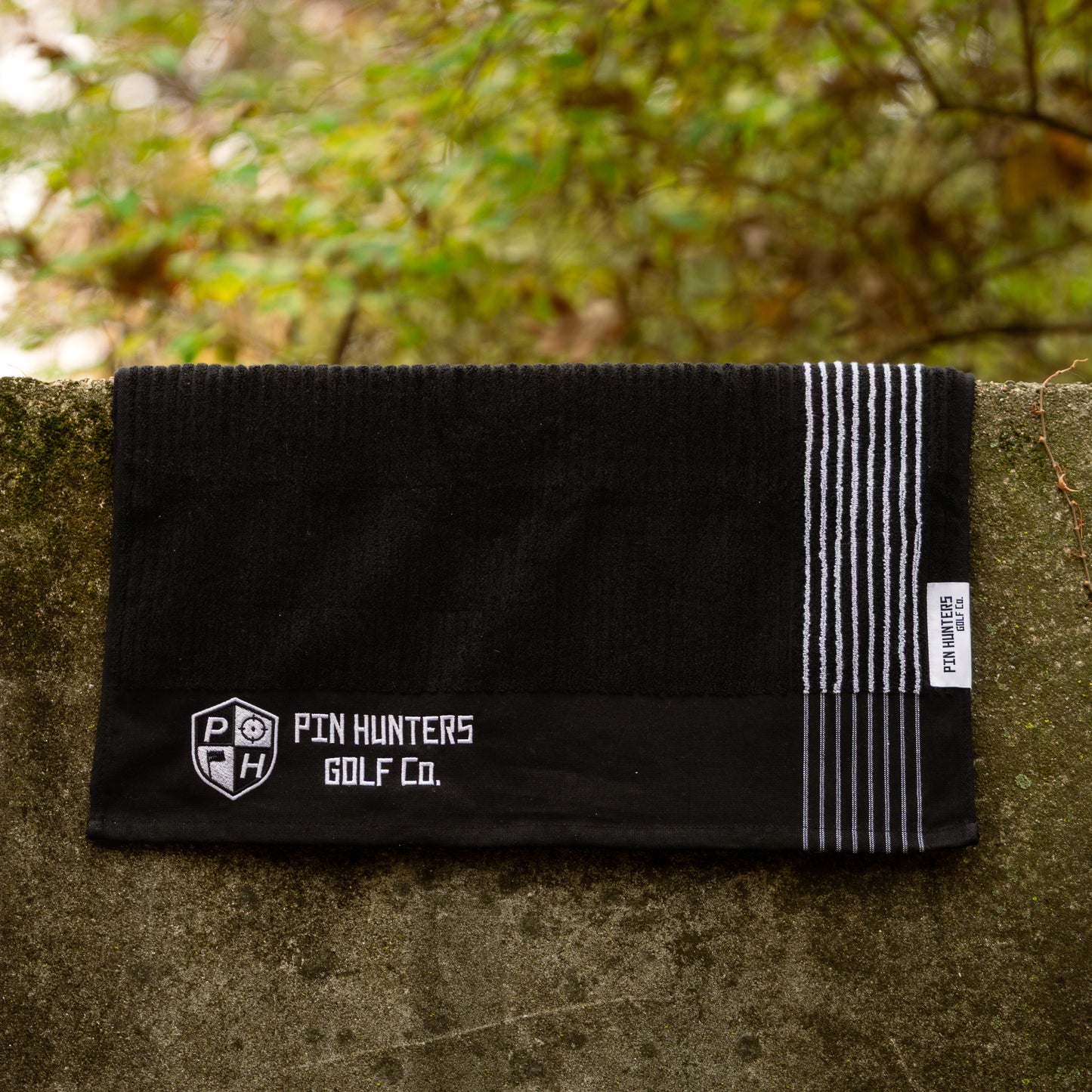 Pin Hunters Golf Co. Looper black cotton golf towel with white stripes hanging on an outdoor stone wall.