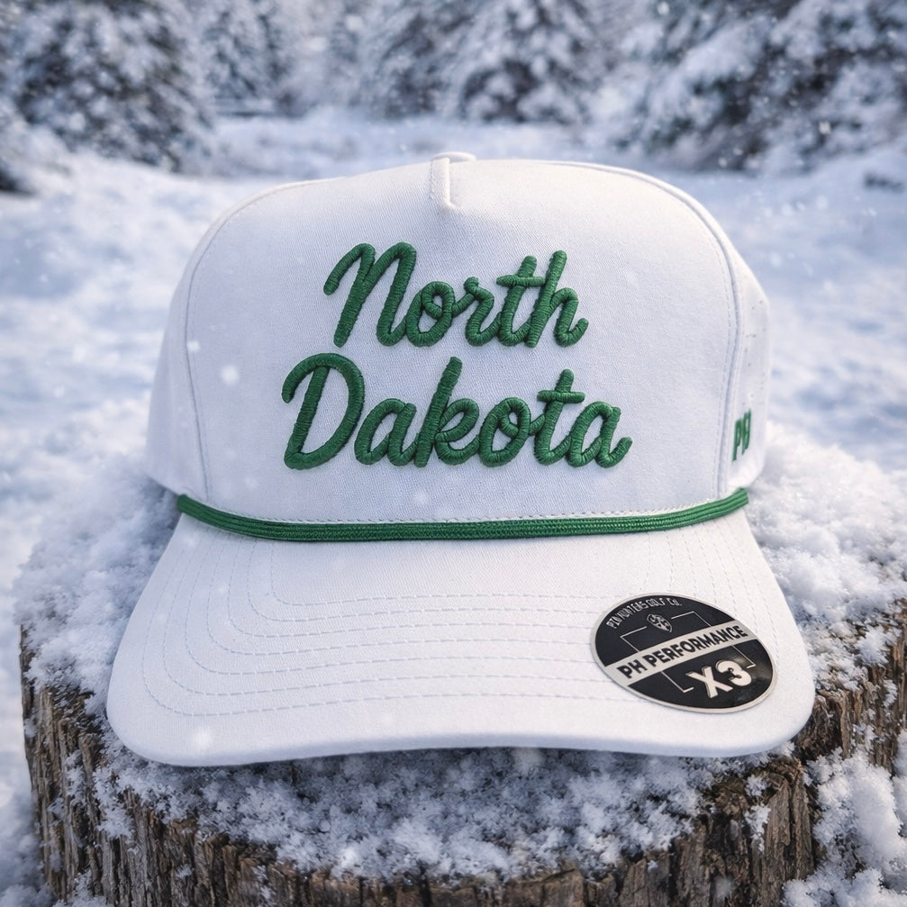 White North Dakota Performance Hat by Pin Hunters Golf Co. with green script embroidery and matching rope detail displayed on a snowy tree stump.