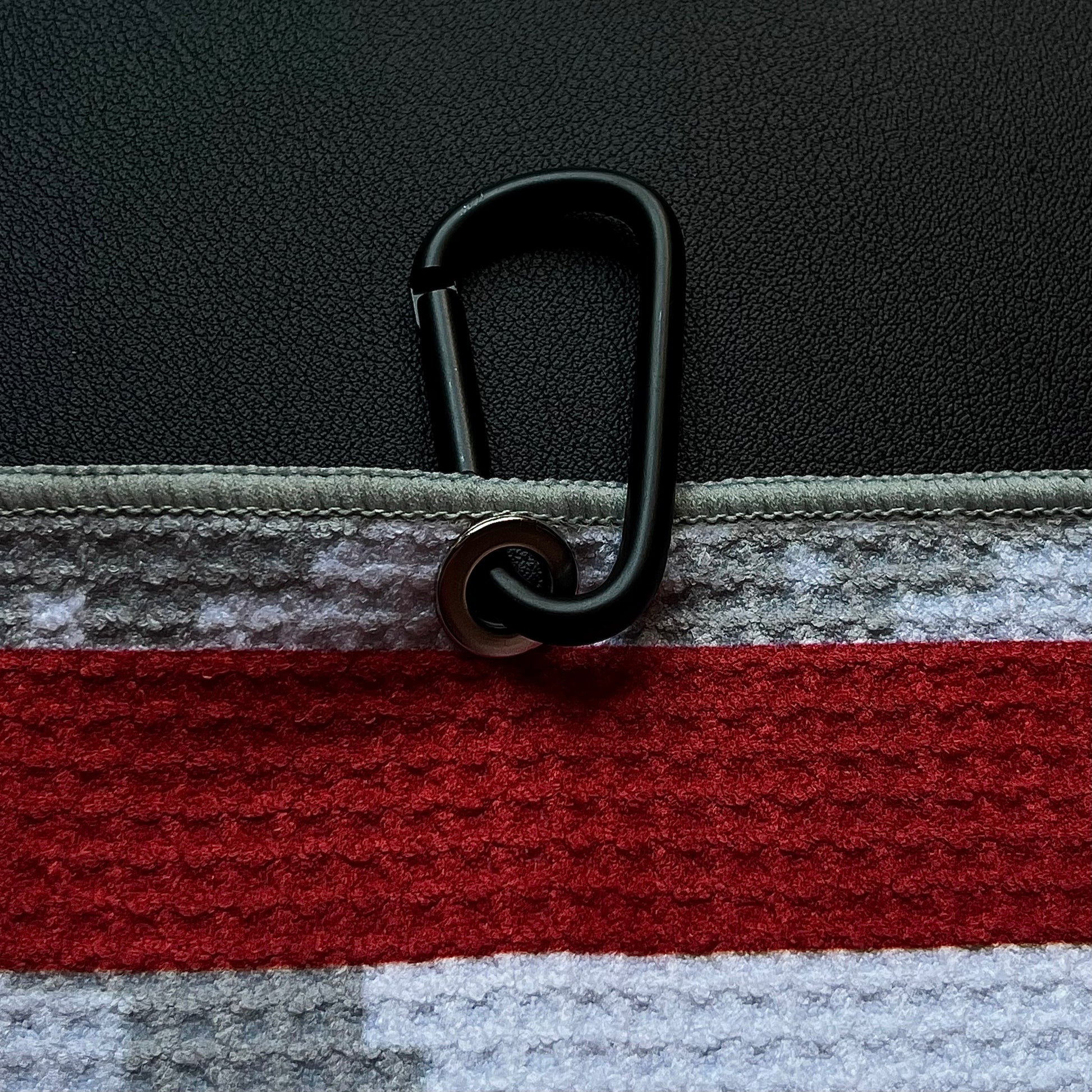 Close up of a black carabiner clipped to an American flag golf towel with red stripe, gray digital camo, and microfiber texture.