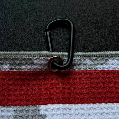 Close up of a black carabiner clipped to an American flag golf towel with red stripe, gray digital camo, and microfiber texture.