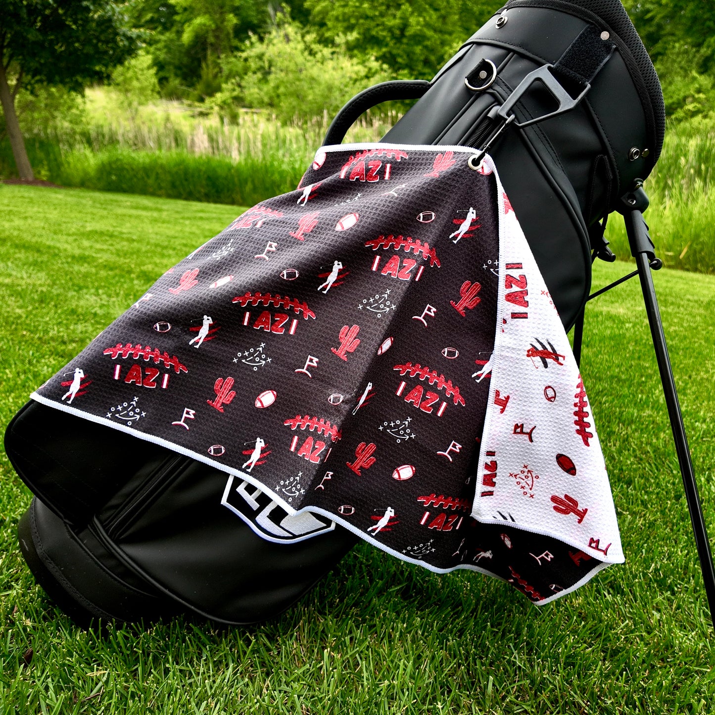 Arizona Cardinals black golf towel hanging on a black golf bag with red AZ football graphics, cactus icons, golfers, footballs, playbook, and white reverse side.