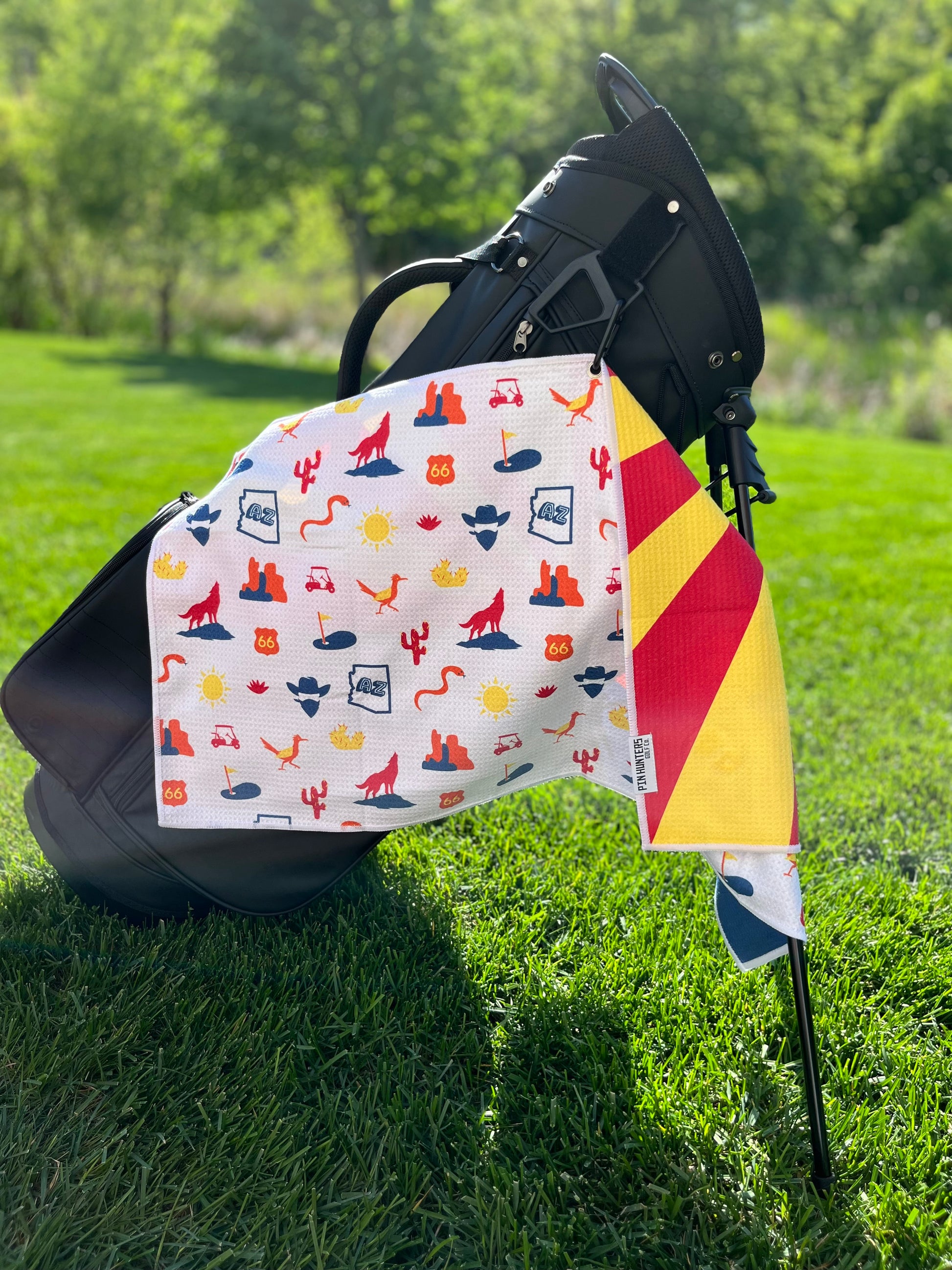 Arizona flag golf towel hanging from a black golf bag, showing the white back pattern with Arizona desert icons, Route 66 signs, cactus, golf carts, golf flags, and red and yellow flag edge.