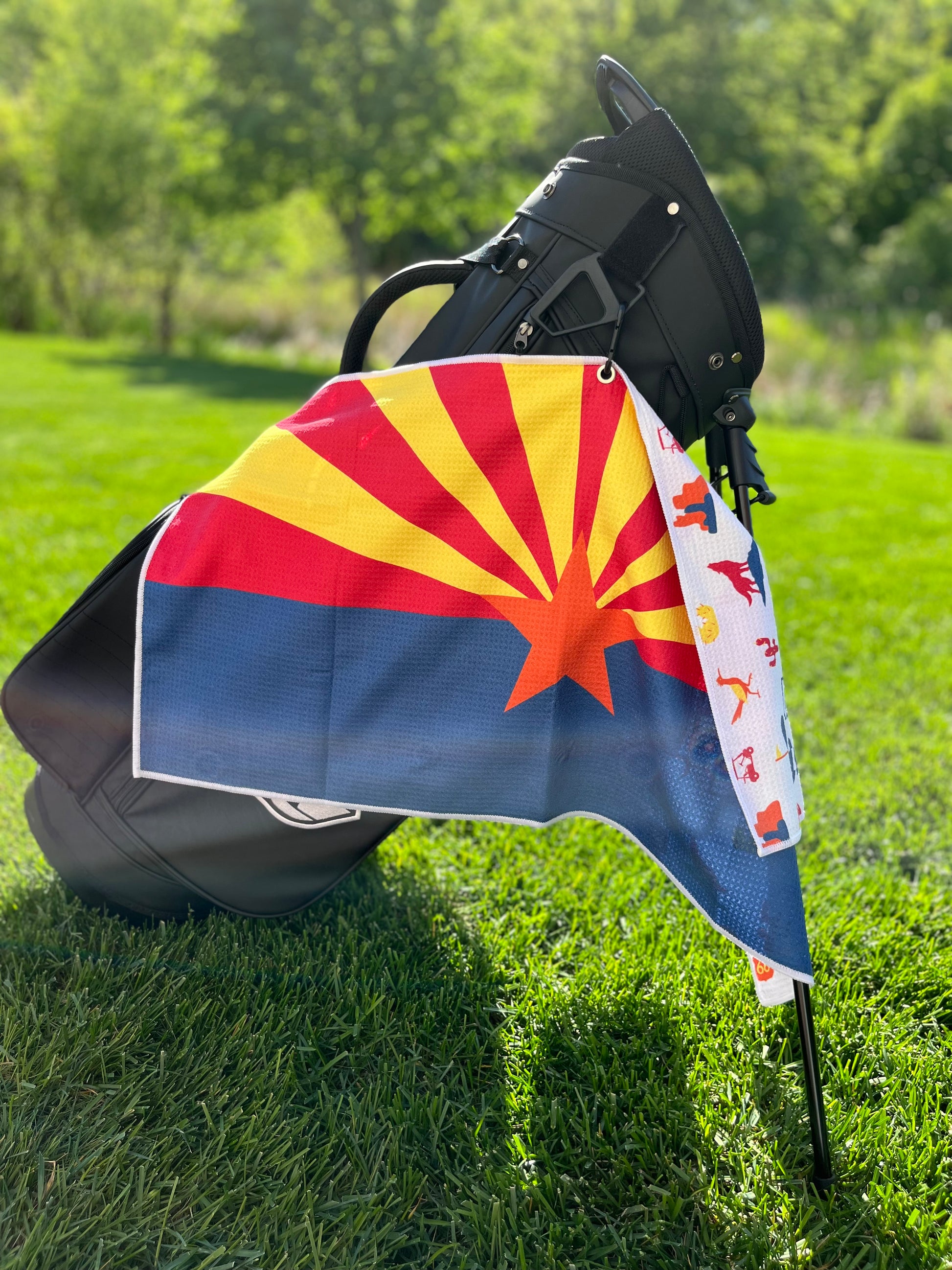 Arizona flag golf towel hanging from a black golf bag with the red and yellow Arizona sunrise design, orange star, and blue lower section.