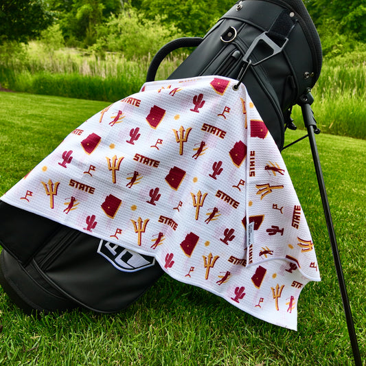 Arizona State Sun Devils white golf towel hanging on a black golf bag with maroon and gold pitchforks, Arizona outlines, cactus icons, sun graphics, golfers, and golf flags.