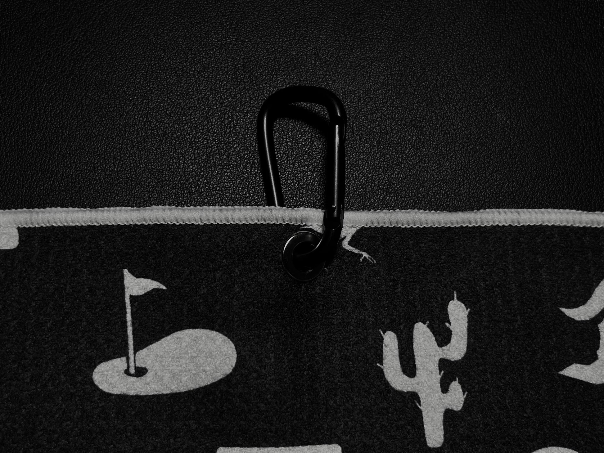 Close up of a black Arizona golf towel clipped with a black carabiner, showing white desert icons, cactus, golf flag, and towel edge detail.
