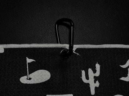 Close up of a black Arizona golf towel clipped with a black carabiner, showing white desert icons, cactus, golf flag, and towel edge detail.
