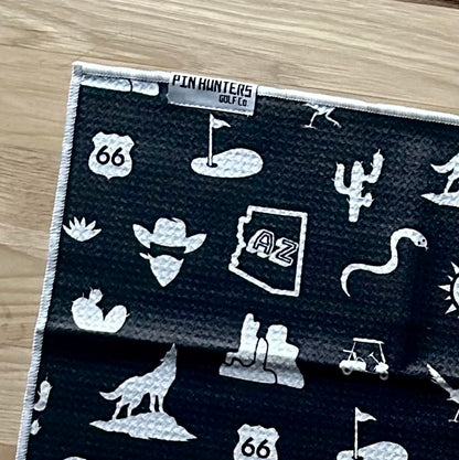 Close up of a black Arizona golf towel with Pin Hunters label, AZ state icon, cactus, Route 66, golf flag, and desert graphics.