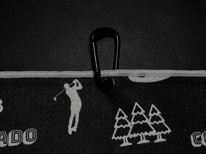 Close up of a black Colorado golf towel with carabiner clip, white golfer, pine tree, mountain, Colorado text, and golf graphics.