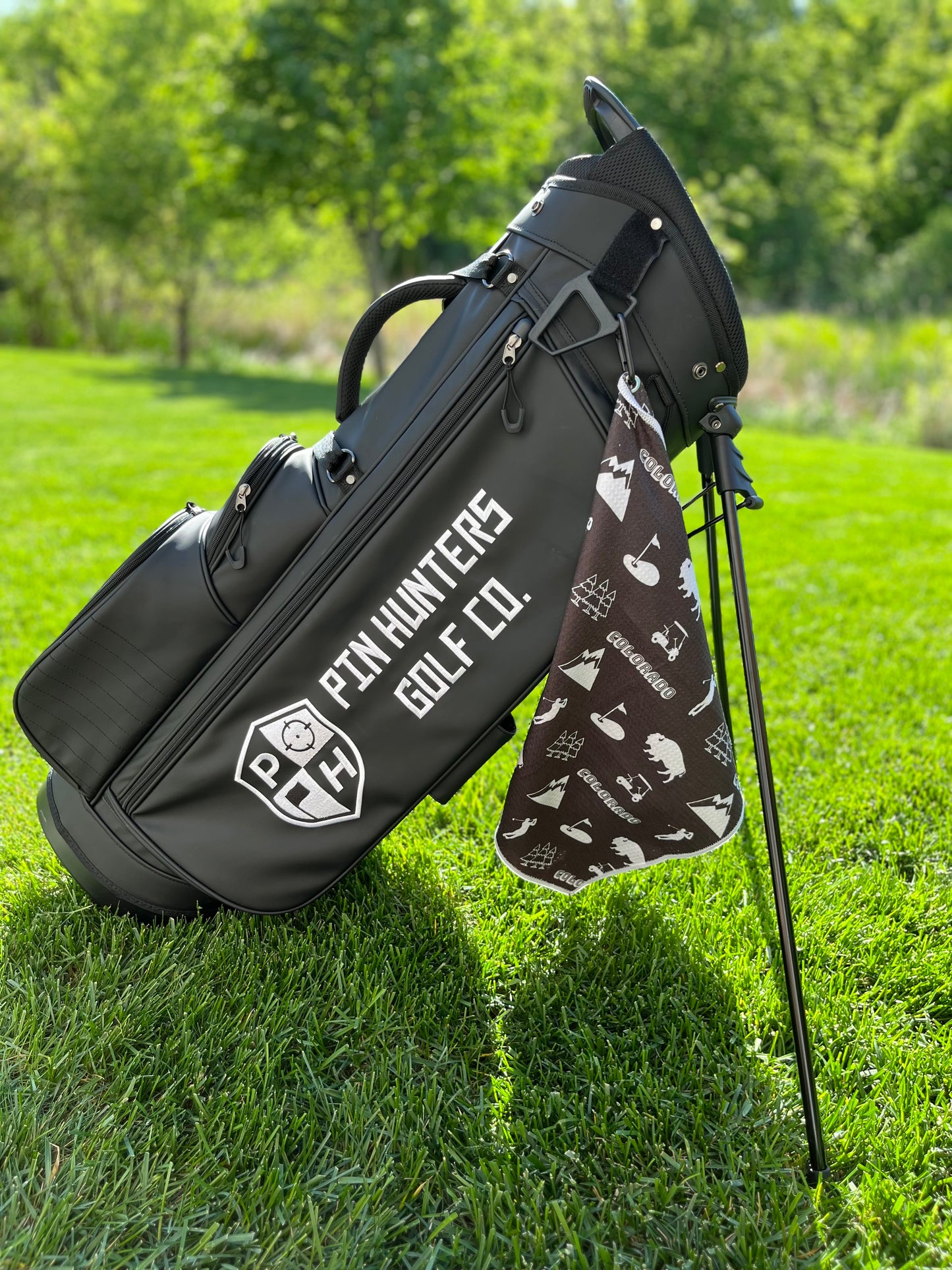 Black Colorado themed golf towel hanging from a black Pin Hunters golf bag with white Colorado, mountain, bison, golfer, and golf icons.