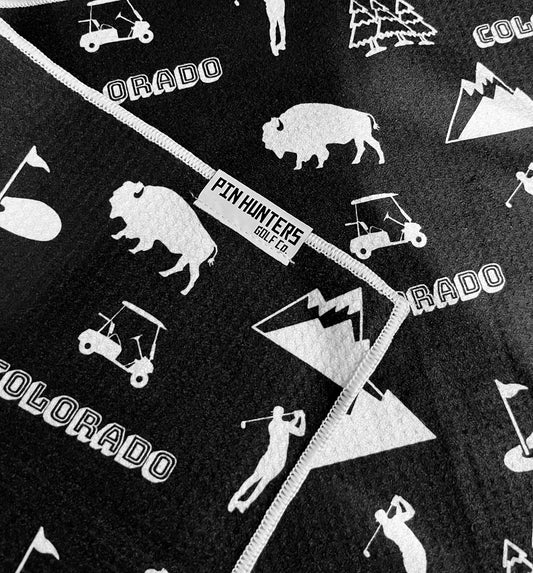 Close up of a black Colorado golf towel with white mountain, bison, golf cart, golfer, and Pin Hunters label details.