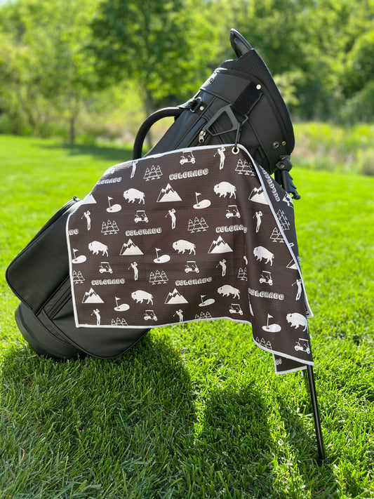 Black Colorado golf towel on a black golf bag with white mountain, bison, golf cart, golfer, pine tree, and flag icons.