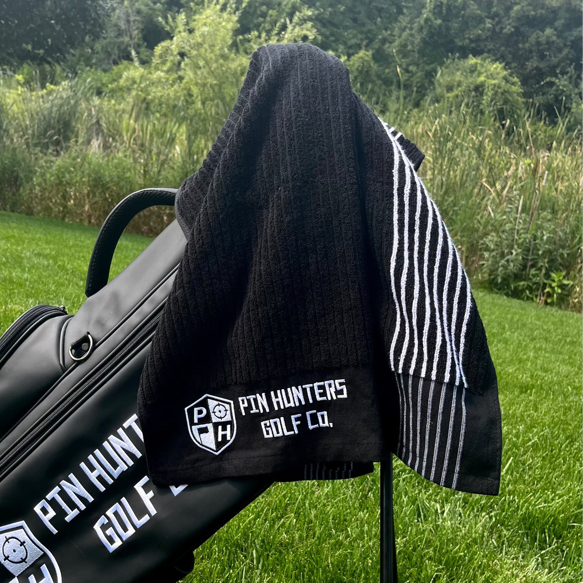 Pin Hunters Golf Co. Looper black cotton golf towel draped over a black golf bag outdoors on the course.