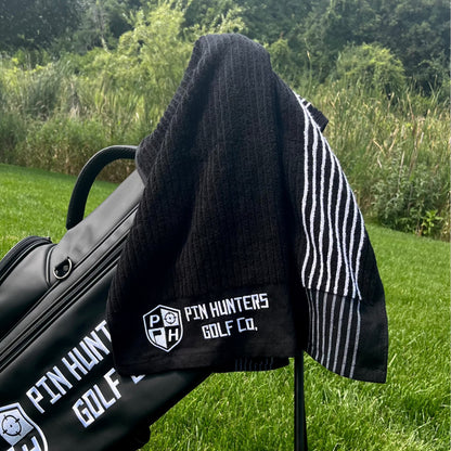 Pin Hunters Golf Co. Looper black cotton golf towel draped over a black golf bag outdoors on the course.