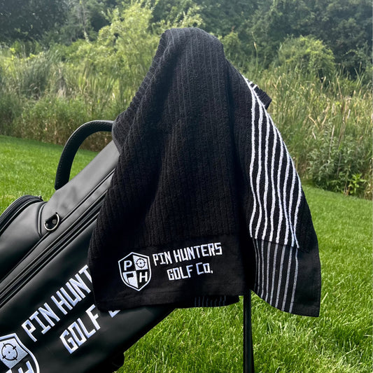 Pin Hunters Golf Co. Looper black cotton golf towel draped over a black golf bag outdoors on the course.