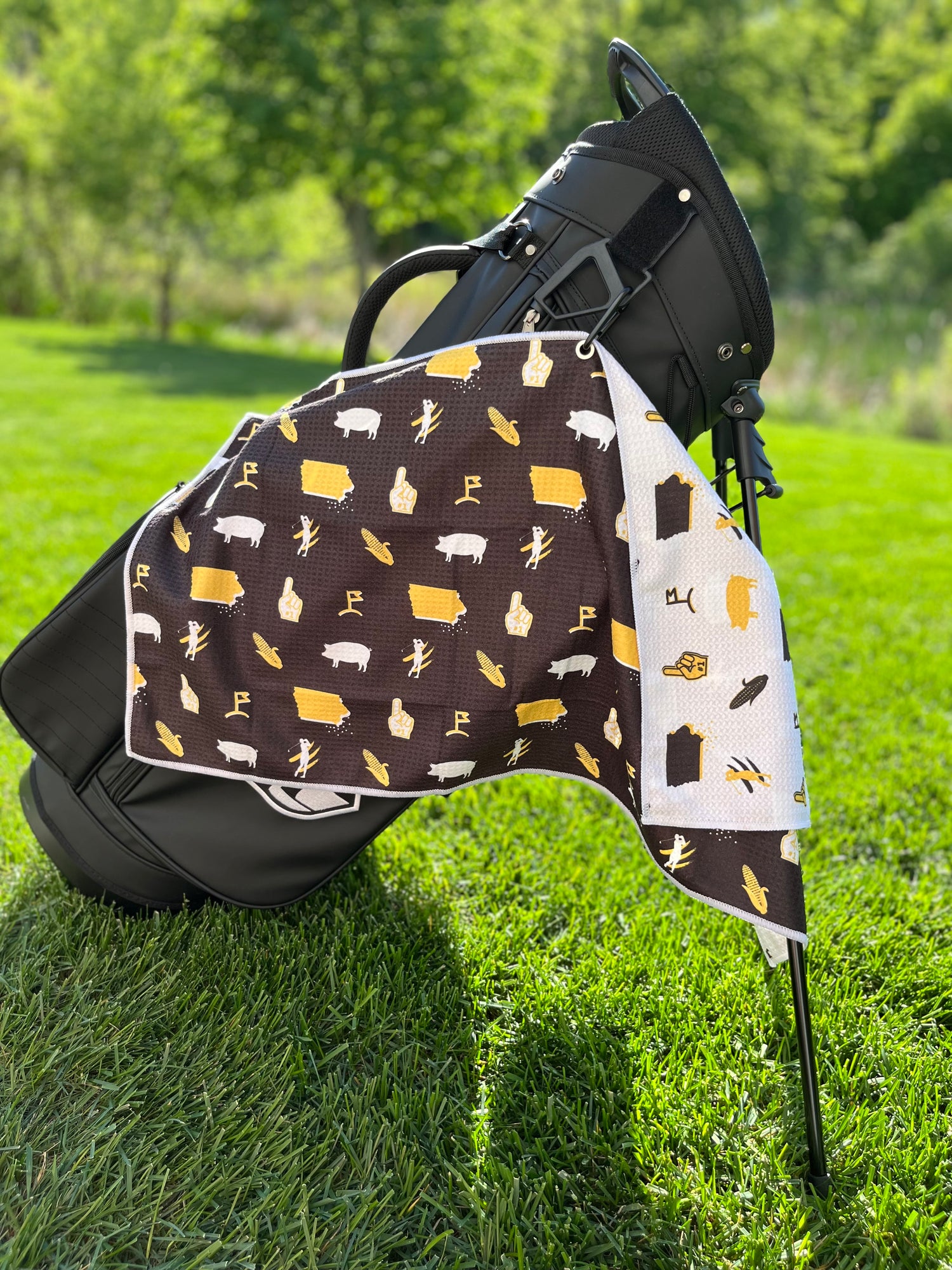 Black and gold Iowa Hawkeyes inspired golf towel hanging from a black golf bag with Iowa state, corn, pig, foam finger, and golf flag graphics.