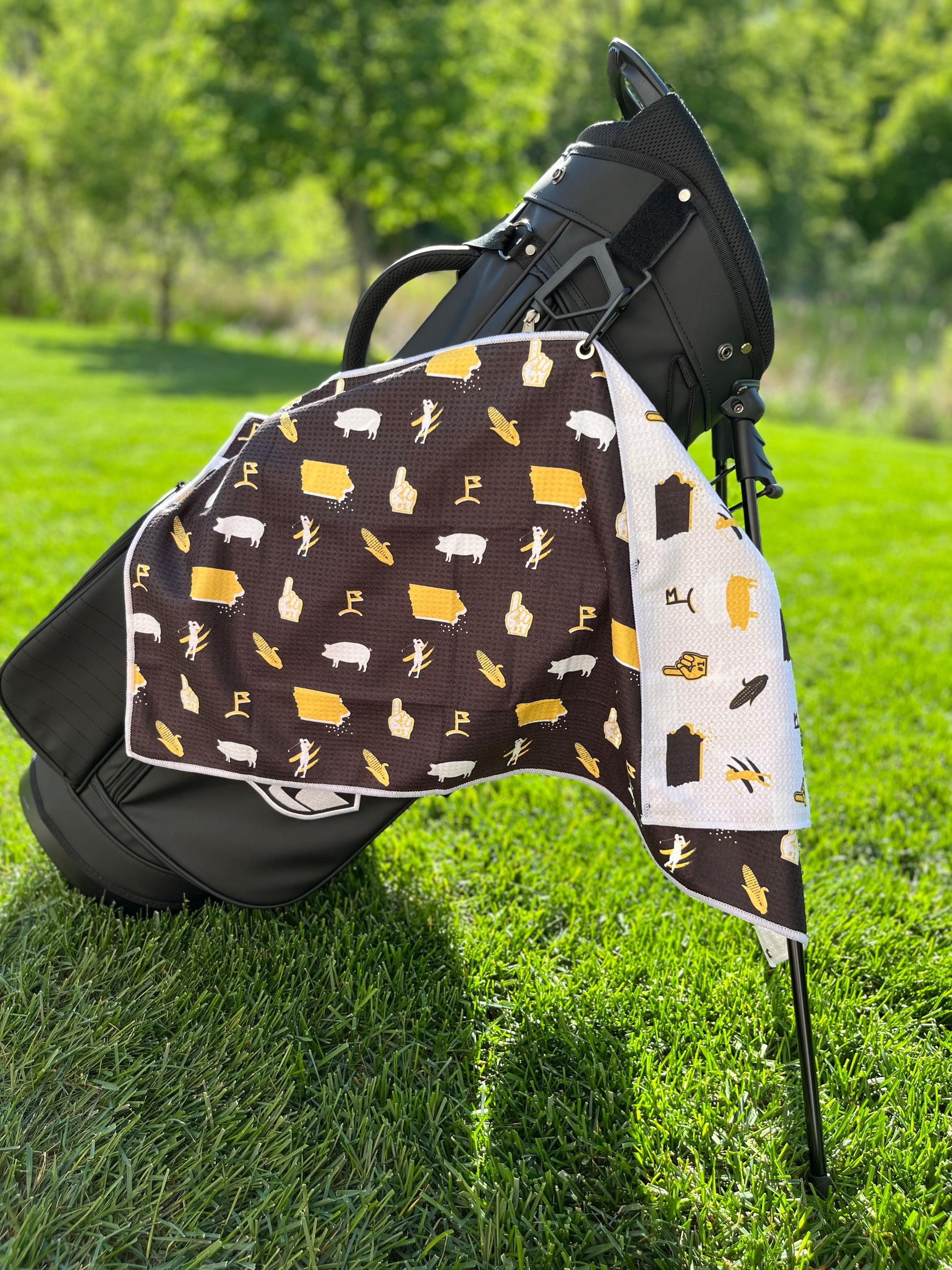 Black and gold Iowa Hawkeyes inspired golf towel hanging from a black golf bag with Iowa state, corn, pig, foam finger, and golf flag graphics.
