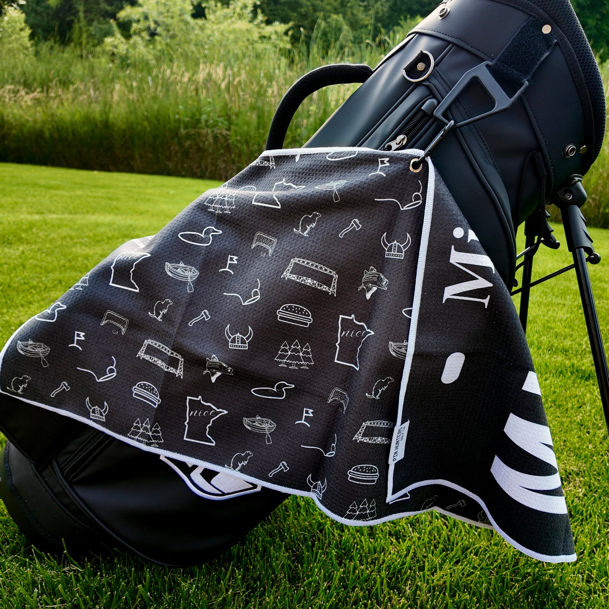 Black Minnesota license plate golf towel hanging on a black golf bag with white Minnesota icons, loons, helmet, bridge, pine trees, golf flags, and state graphics.