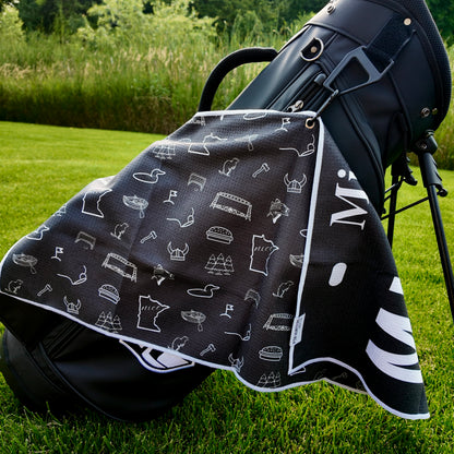 Black Minnesota license plate golf towel hanging on a black golf bag with white Minnesota icons, loons, helmet, bridge, pine trees, golf flags, and state graphics.