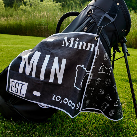 Black Minnesota license plate golf towel hanging on a black golf bag with large white MIN text, Minnesota outline, established date detail, and black-and-white back pattern.
