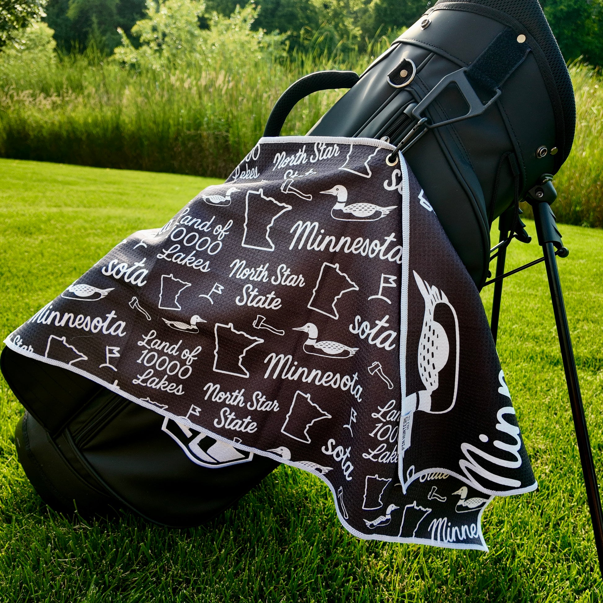Black Minnesota SOTA golf towel hanging from a black golf bag with loons, Minnesota outlines, North Star State text, and golf icons.