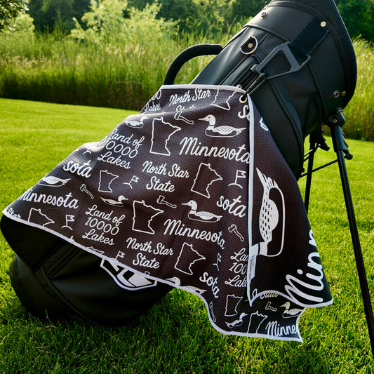 Black Minnesota SOTA golf towel hanging from a black golf bag with loons, Minnesota outlines, North Star State text, and golf icons.