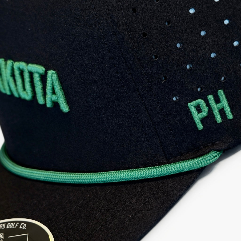 Black North Dakota Performance Hat by Pin Hunters Golf Co. with green embroidery displayed on a snowy tree stump outdoors.