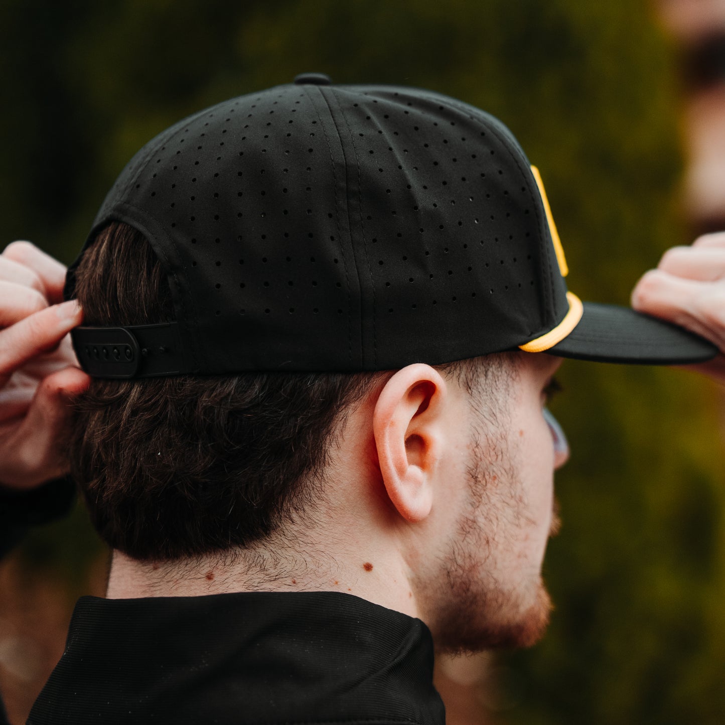 Black performance golf hat back and side view with perforated panels, black snapback closure, and yellow rope detail at front.