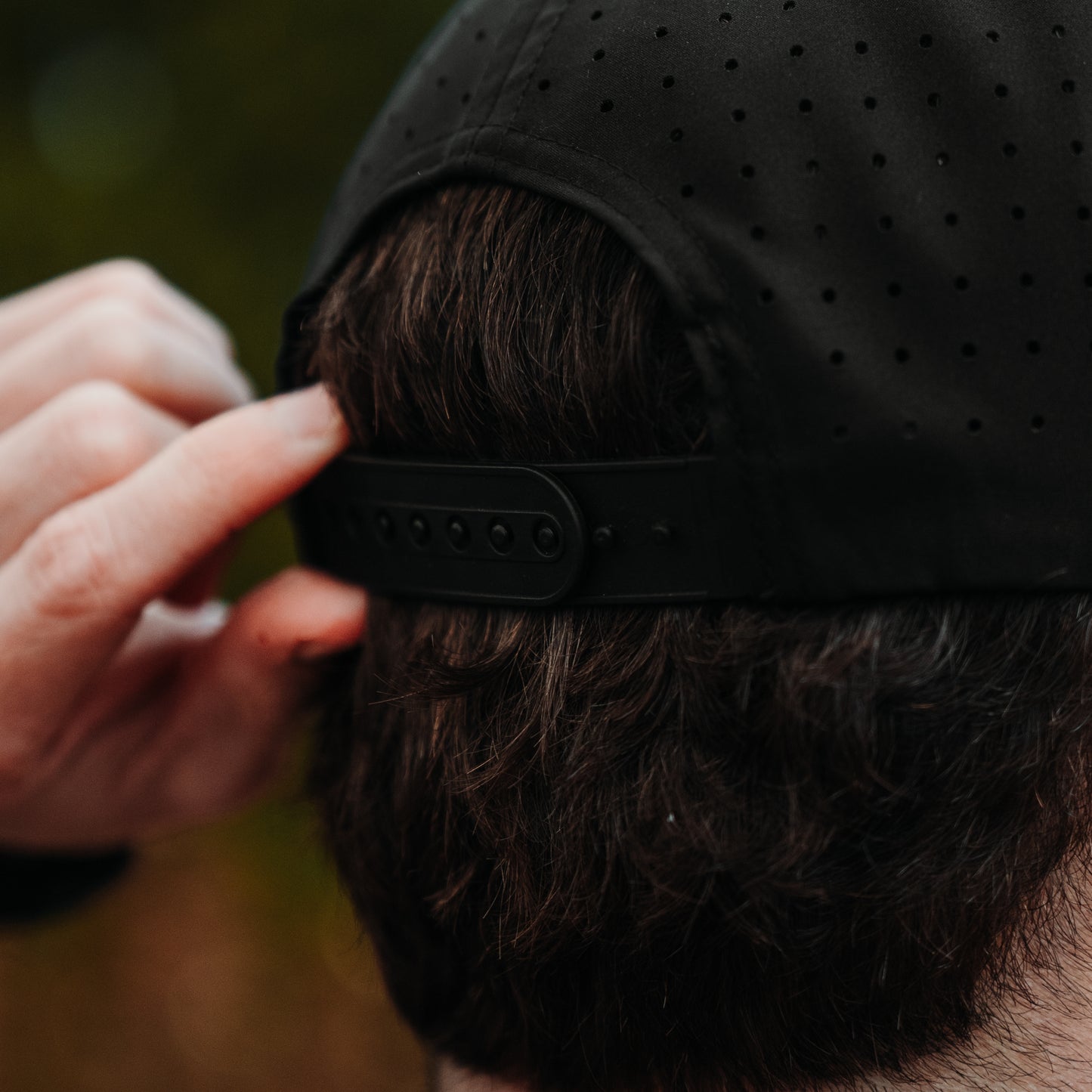 Black performance golf hat back close-up with perforated panels, black snapback closure, and model adjusting the strap.