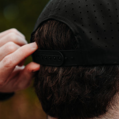 Black performance golf hat back close-up with perforated panels, black snapback closure, and model adjusting the strap.