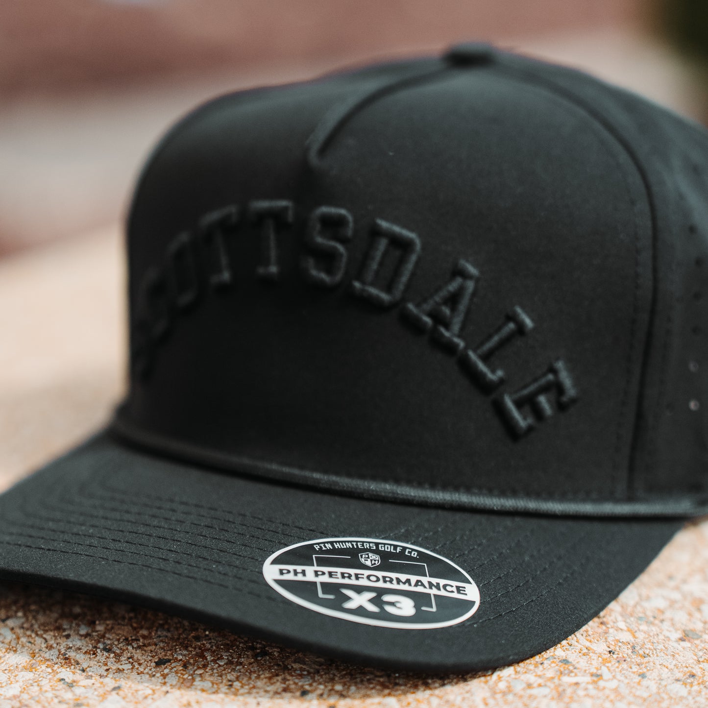 Black Scottsdale tonal performance golf hat close-up with black raised embroidery, matching rope detail, and PH Performance X3 sticker.