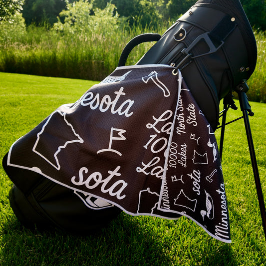 Close up of a black SOTA Minnesota golf towel with white Minnesota outline, loon, Land of 10000 Lakes text, and golf flag graphics.