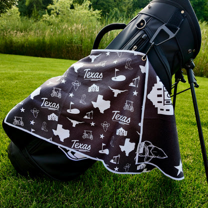 Black Texas themed golf towel hanging from a black golf bag with Texas state, Alamo, longhorn, cowboy boot, oil, and golf icons.