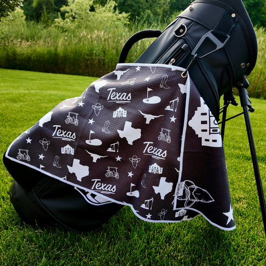 Black Texas themed golf towel hanging from a black golf bag with Texas state, Alamo, longhorn, cowboy boot, oil, and golf icons.