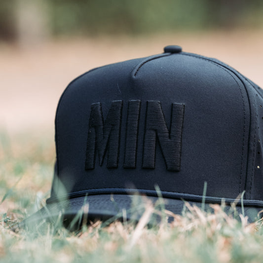 Black tonal MIN performance golf hat front close-up with raised MIN embroidery, matching rope detail, flat brim, and low-angle grass background.