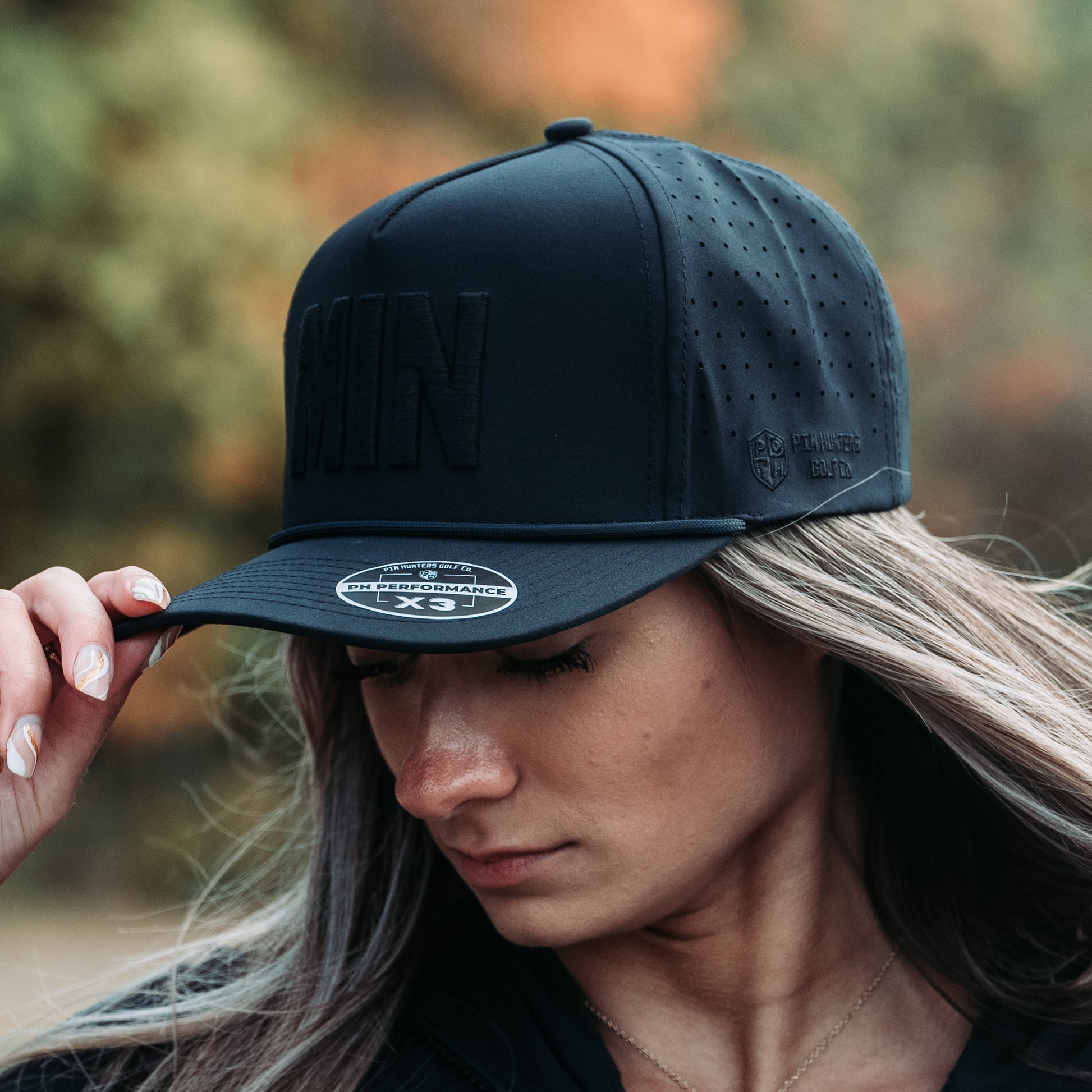 Black tonal MIN performance golf hat on model with raised black embroidery, side logo, perforated panels, rope detail, and PH Performance X3 sticker.