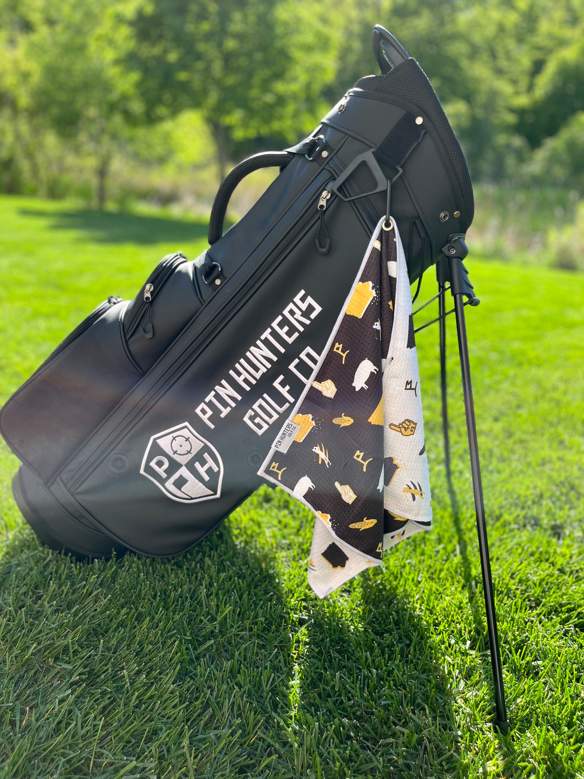 Black, white, and gold Iowa Hawkeyes inspired golf towel hanging from a black Pin Hunters golf bag with Iowa, corn, pig, and golf icons.