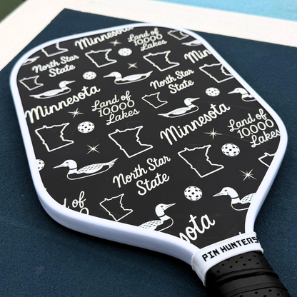 Black and white Minnesota pickleball paddle close-up with loon icons, state outlines, North Star State text, and Land of 10,000 Lakes graphics.