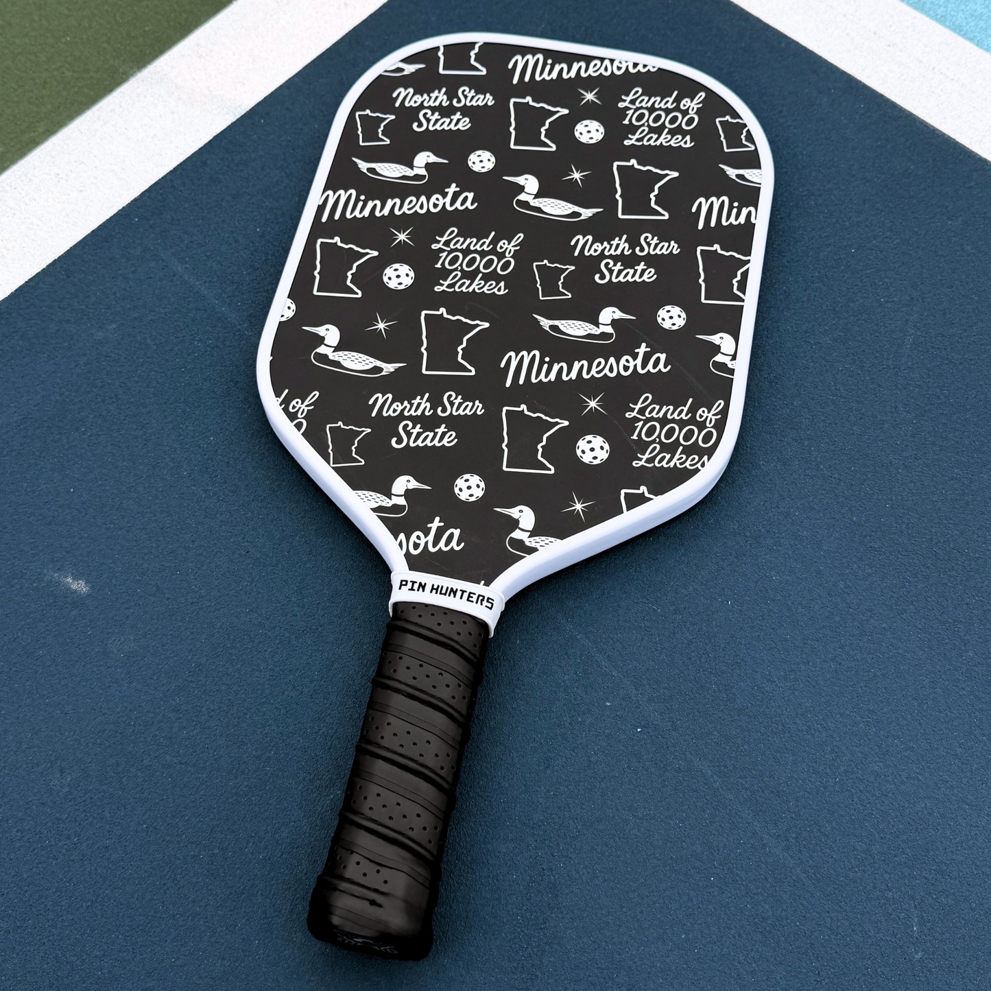 Black and white Minnesota pickleball paddle on court with loon icons, state outlines, North Star State text, and Land of 10,000 Lakes pattern.