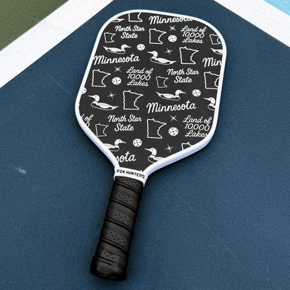 Black and white Minnesota pickleball paddle on court with loon icons, state outlines, North Star State text, and Land of 10,000 Lakes pattern.