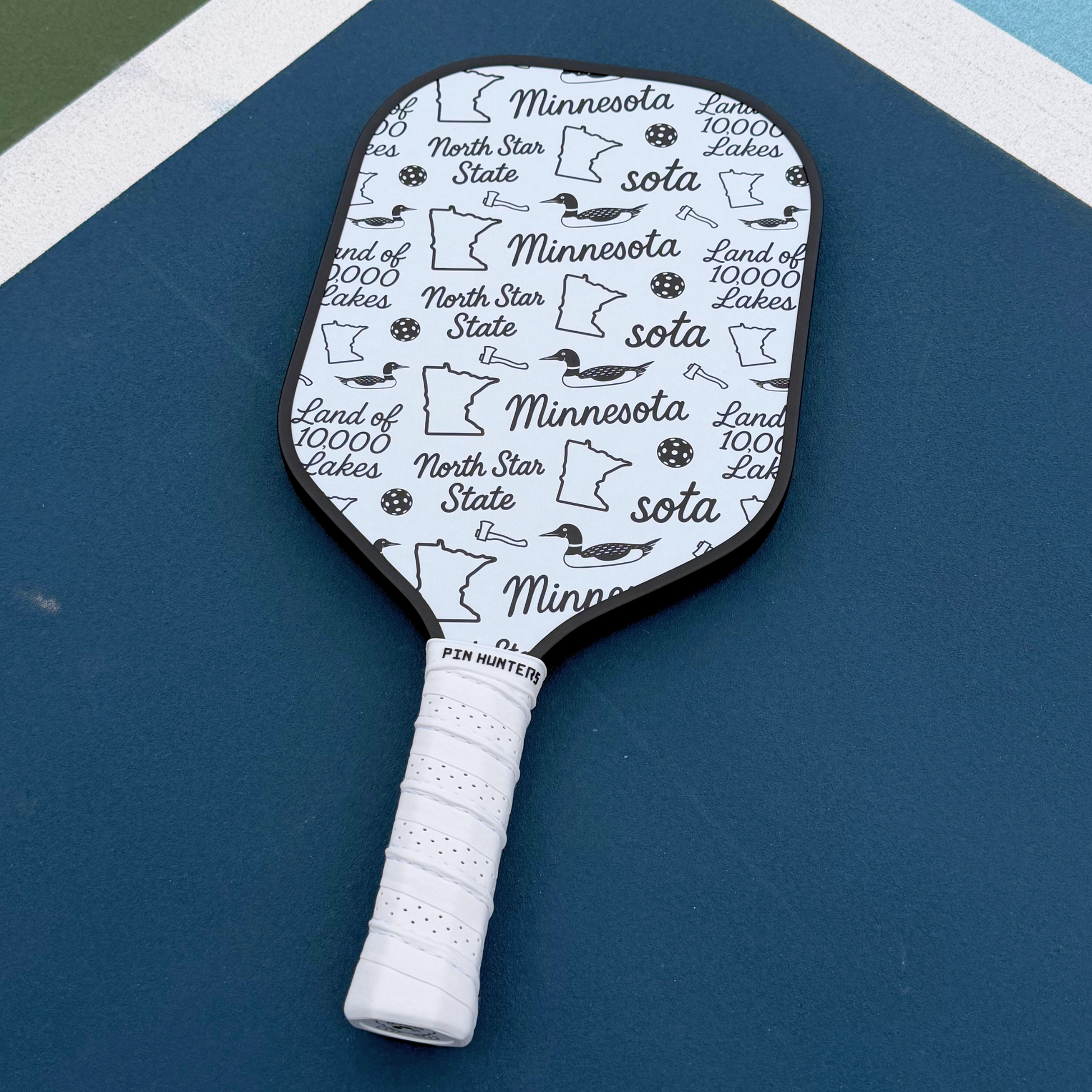 Black and white Minnesota pickleball paddle on court with state outline, loons, North Star State, Land of 10,000 Lakes, and pickleball icons.